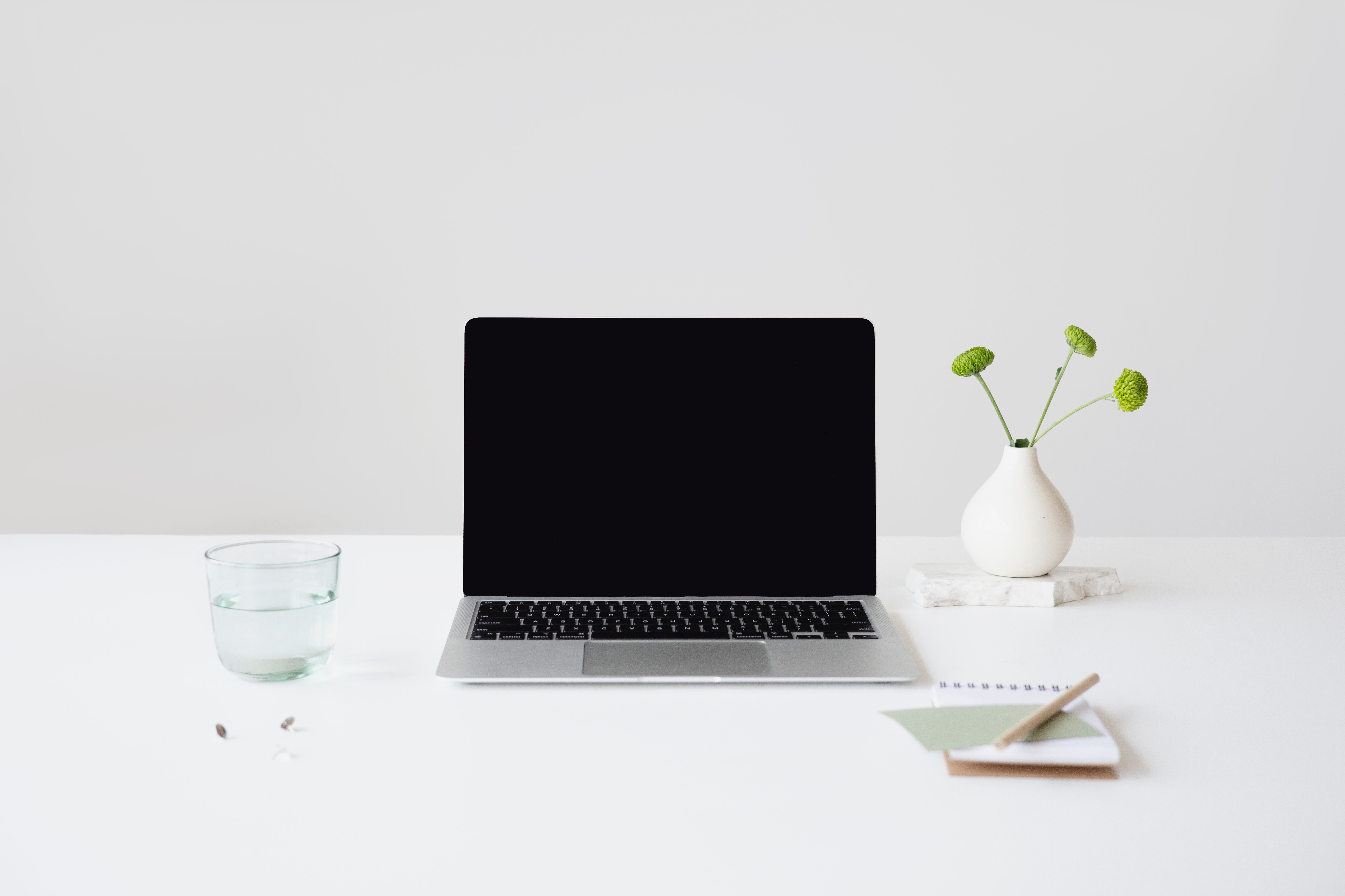 Photo of glass of water, open MacBook with screen off, plant and notepad on white table. 